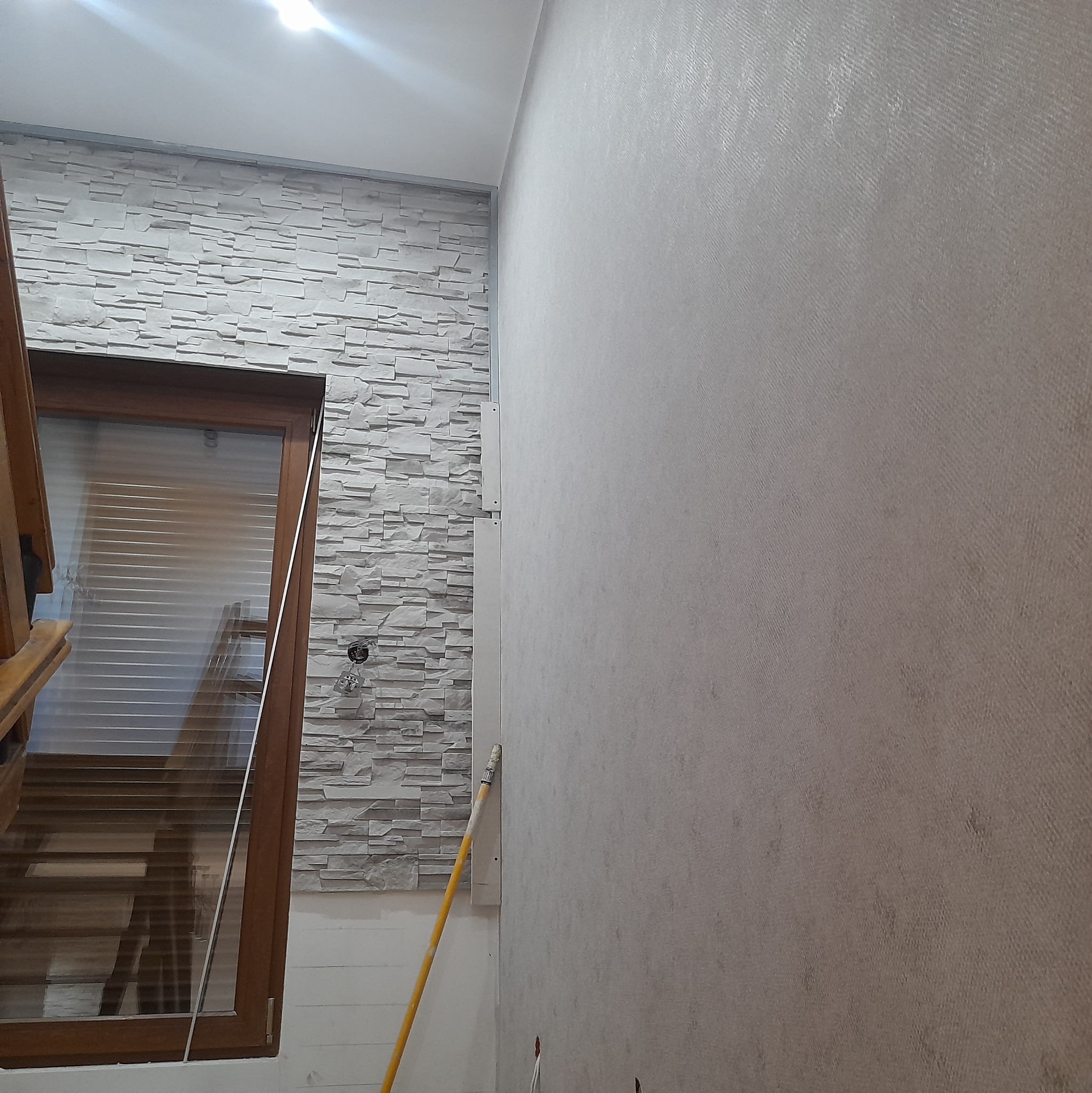 Fragment wnętrza z dekoracyjną ścianą z imitacji kamienia, drewnianą ramą okienną i pomalowaną ścianą z widocznym wałkiem malarskim.