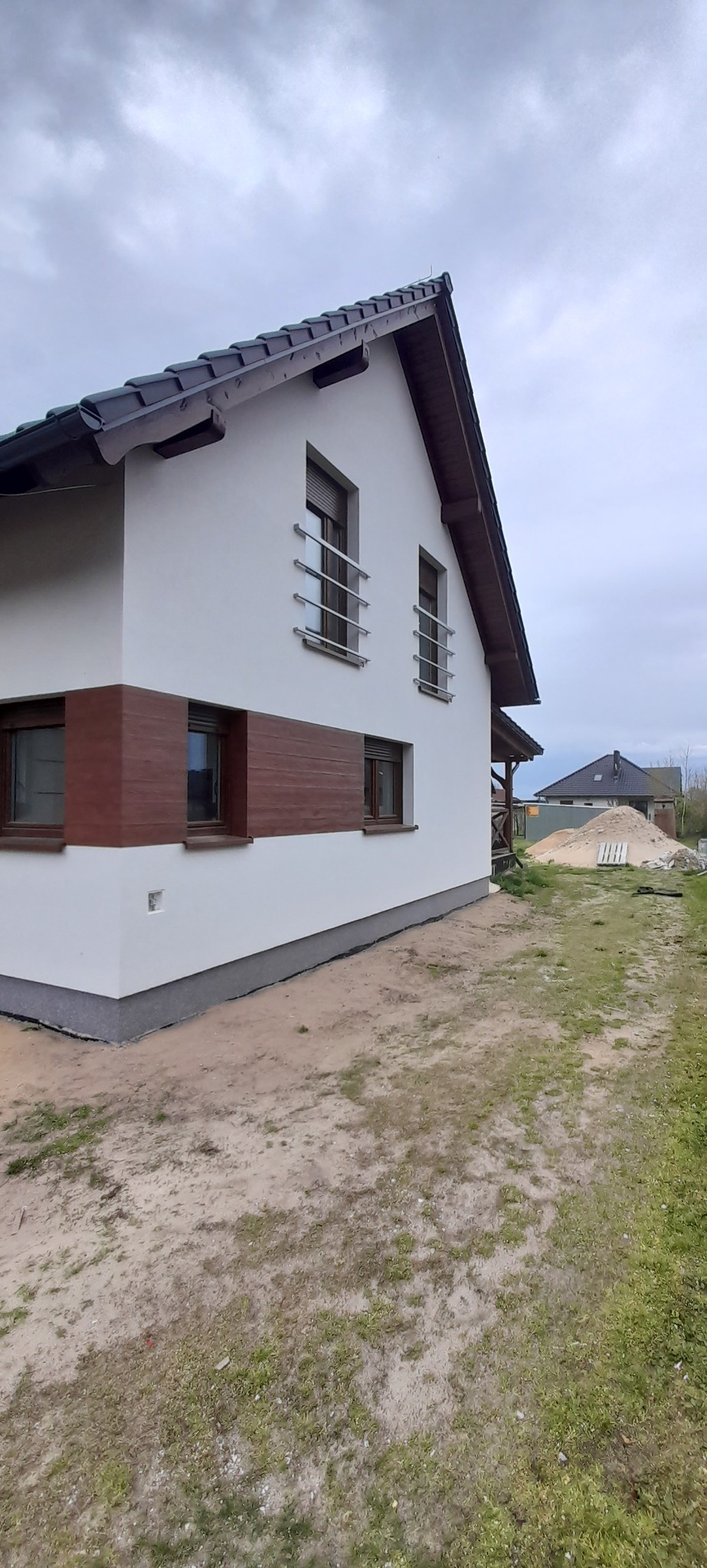 Nowoczesny dom jednorodzinny z białymi ścianami, brązowymi akcentami i ciemną dachówką, widoczny z boku na tle pochmurnego nieba i placu budowy.