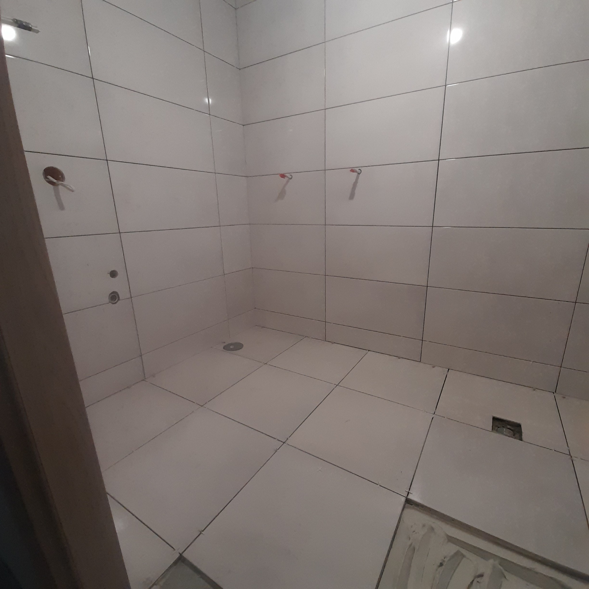 Łazienka w trakcie układania białych płytek ściennych i podłogowych, widoczne otwory na armaturę i odpływ pod prysznicem, krzyżyki dystansowe między płytkami.