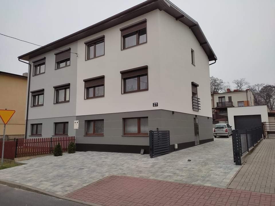 Dwukondygnacyjny dom z elewacją w dwóch odcieniach szarości i brązowymi roletami, numer domu 27, widok z poziomu ulicy w pochmurny dzień.