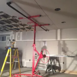 Czerwony podnośnik do płyt gipsowych podtrzymuje zamontowaną płytę na suficie, widoczny fragment drewnianej konstrukcji sufitu podwieszanego, drabina i narzędzia w tle.
