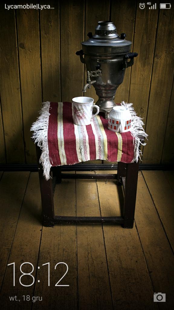 Zdjęcie ekranu smartfona z widocznym samowarem, filiżanką i cukiernicą na małym stoliku przykrytym tkaniną w paski, ustawionymi na tle drewnianej ściany i podłogi; na pierwszym planie widoczny...