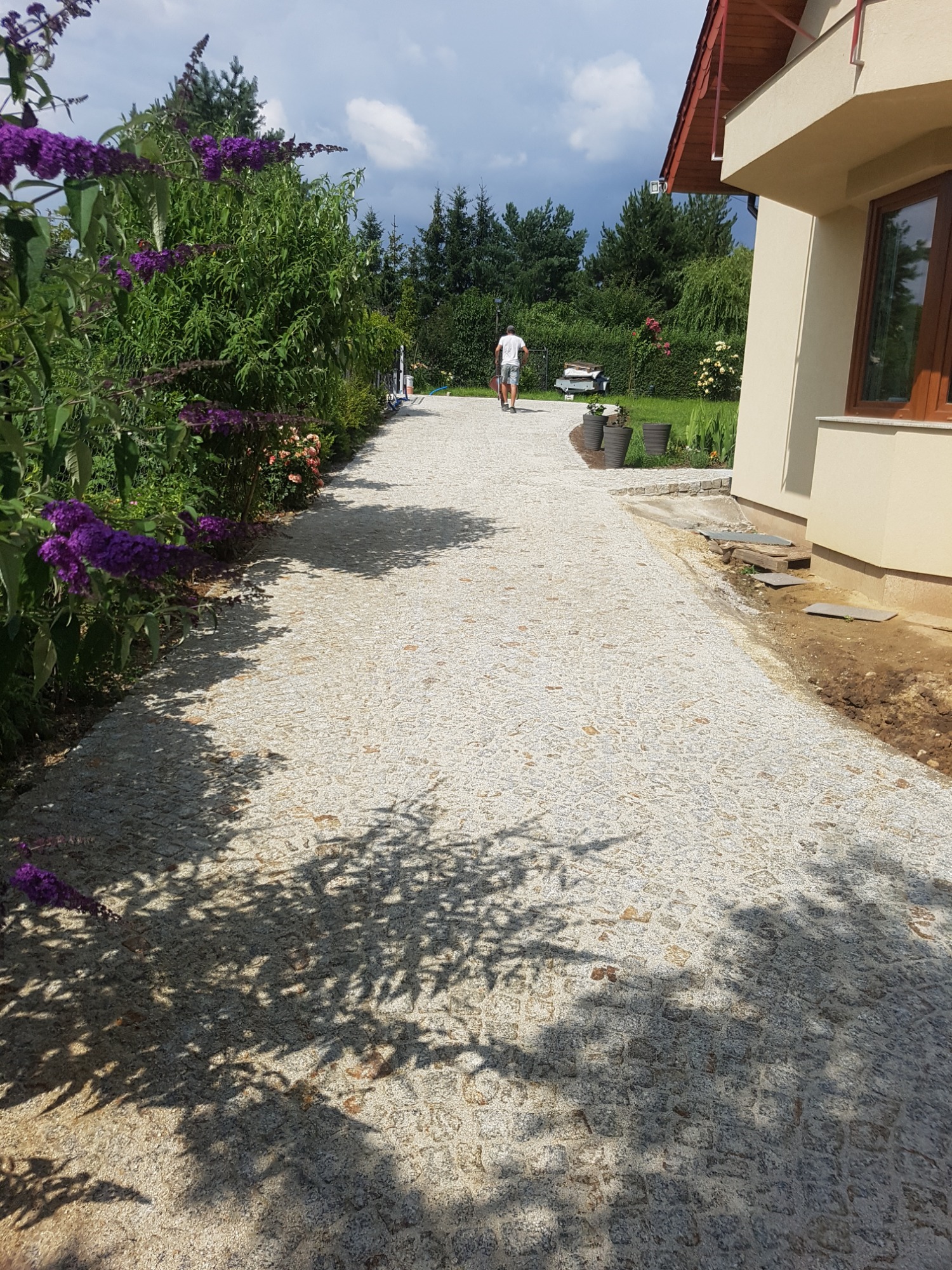 Długa, jasna ścieżka z drobnej kostki prowadząca wzdłuż domu i ogrodu, z widocznymi cieniami drzew i mężczyzną z taczką w oddali.