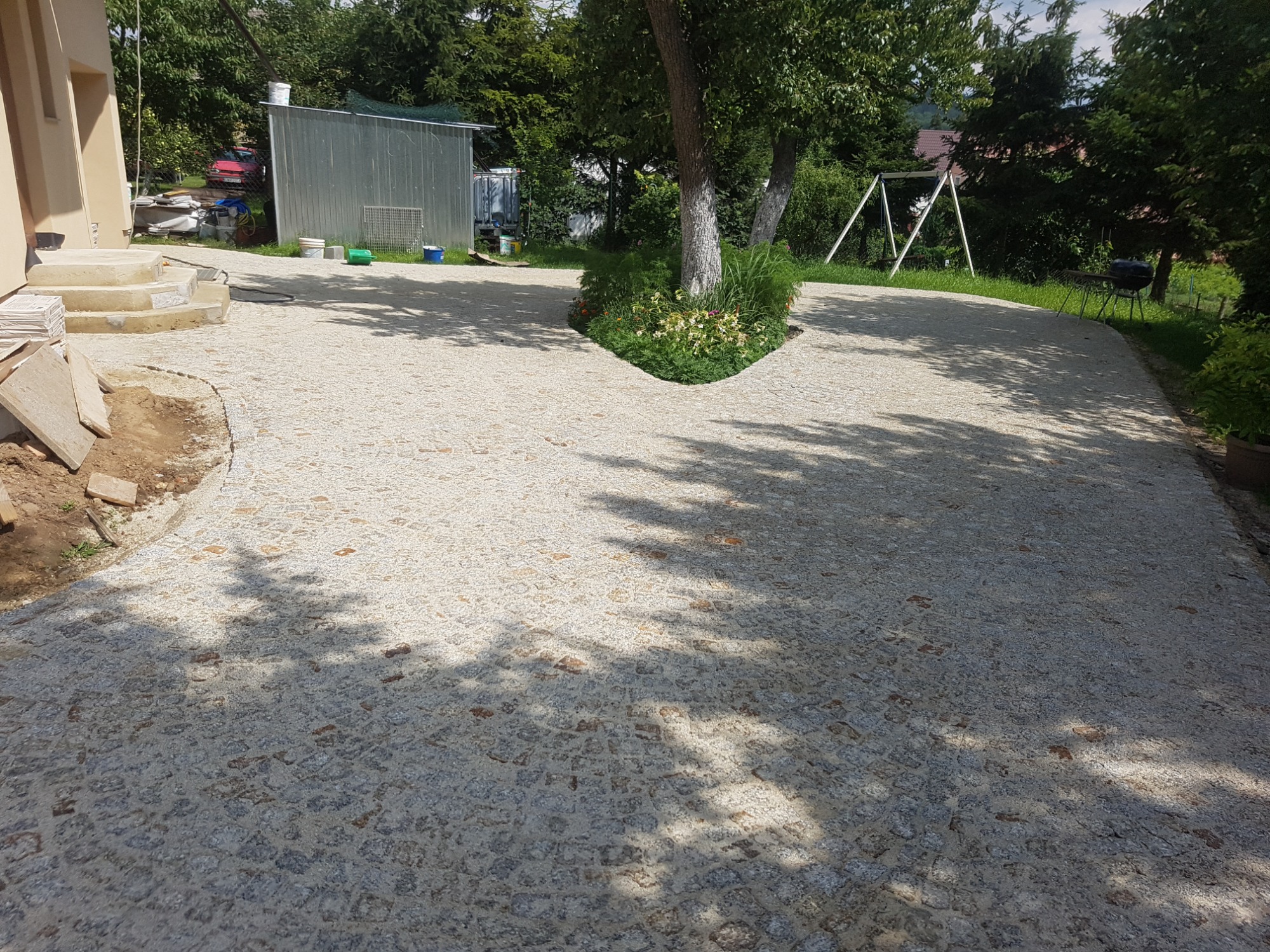 Szeroka aleja z jasnoszarej kostki brukowej rozwidlająca się wokół drzewa z rabatą kwiatową, w tle budynek gospodarczy i huśtawka ogrodowa.