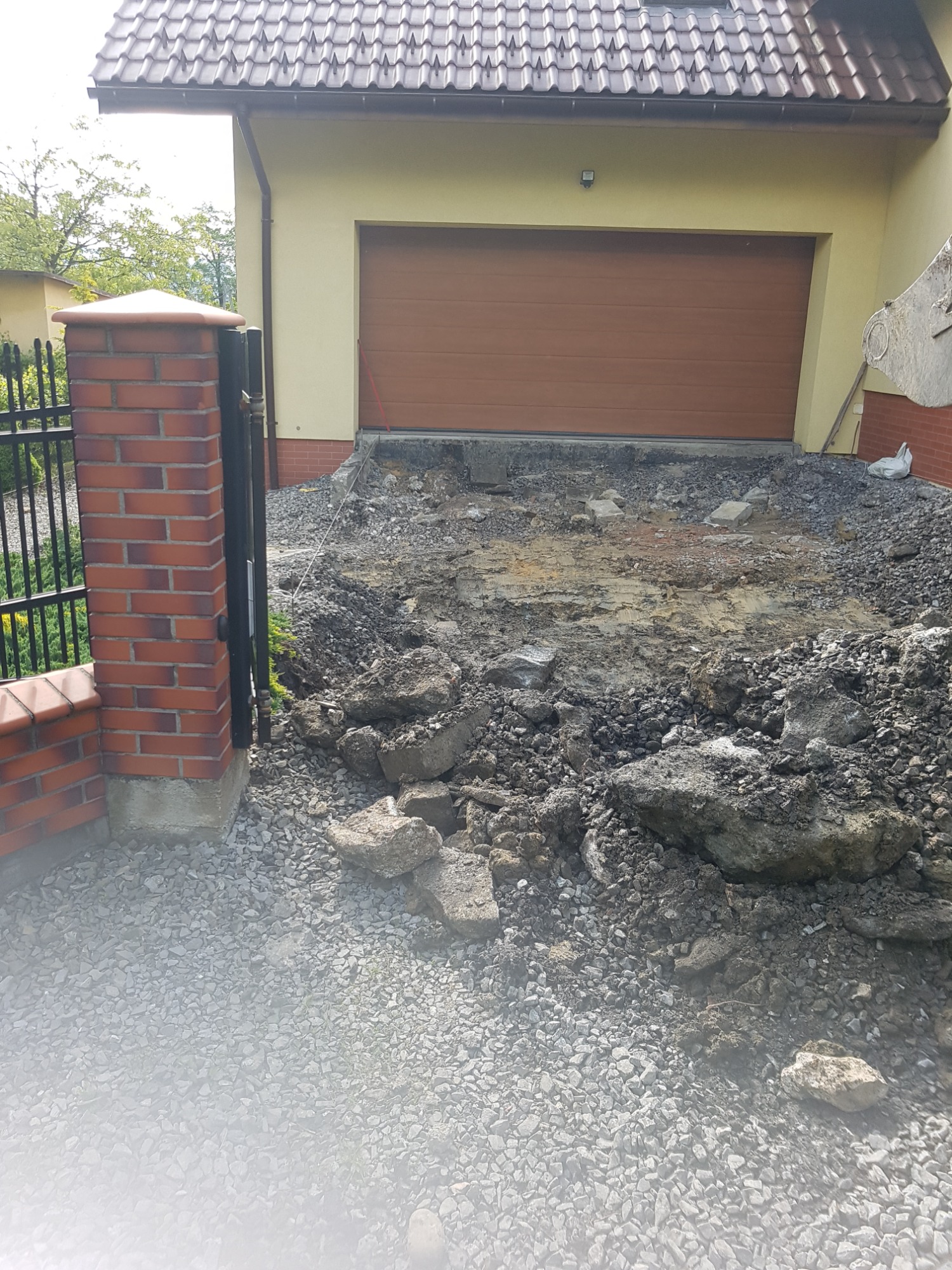 Teren przed garażem w trakcie przygotowania do ułożenia nawierzchni, widoczne gruz, ziemia i podsypka żwirowa.