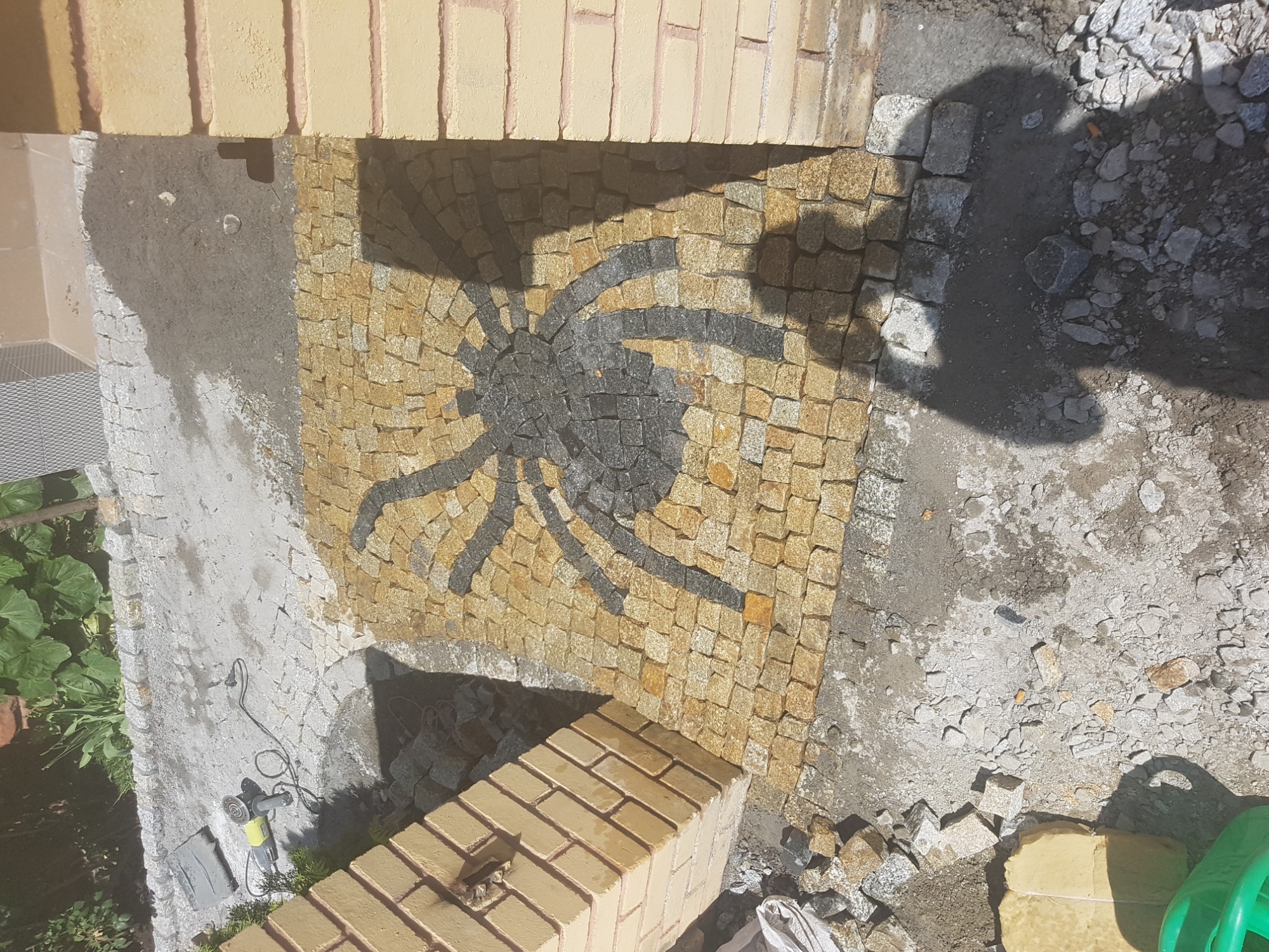 Mozaika z kostki granitowej w kształcie pająka, w trakcie układania, widoczne narzędzia i materiały budowlane.