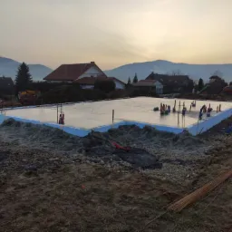 Wylana płyta fundamentowa z niebieskim obrzeżem styropianowym, wystające pręty zbrojeniowe, widok na góry i zabudowania w tle, pora dnia zmierzch.