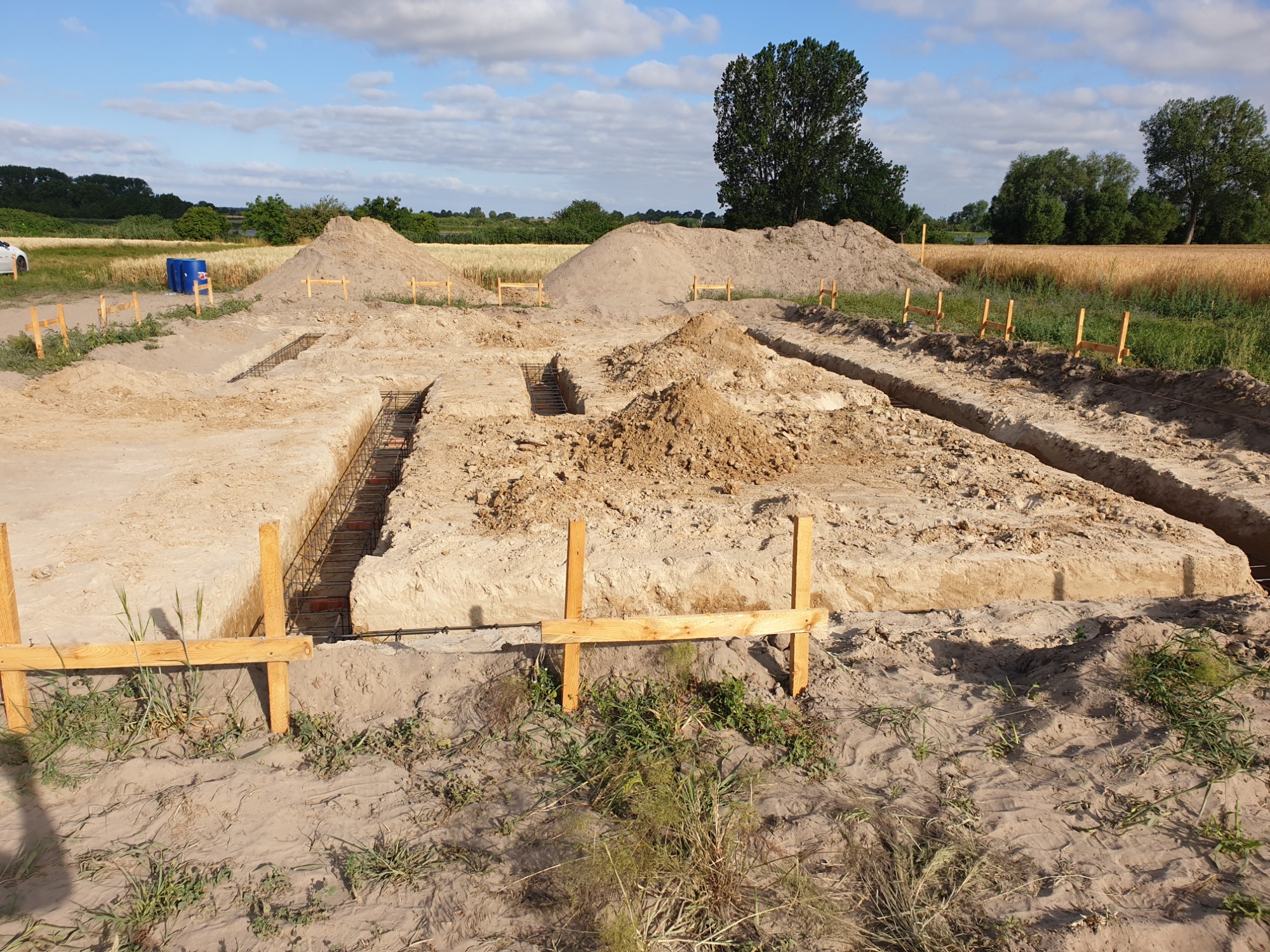Wykopane fundamenty pod budowę domu z widocznym zbrojeniem, drewnianymi szalunkami i stertami ziemi w tle krajobrazu polnego z drzewami i błękitnym niebem.