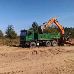 Pomarańczowa koparka ładuje urobek do zielonej wywrotki MAN na placu budowy w słoneczny dzień.