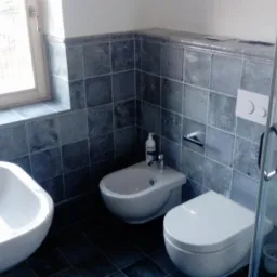 Nowoczesna łazienka z szarymi płytkami, umywalką, bidetem i toaletą w stylu minimalistycznym, kabina prysznicowa z metalowymi uchwytami, okno z widokiem na zewnątrz.