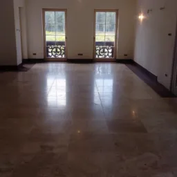 Jasna, polerowana podłoga z płytek imitujących marmur w przestronnym, pustym pokoju z dużymi oknami i niedokończonym kominkiem, odbijająca światło.