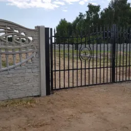 Czarna, metalowa brama wjazdowa z ozdobnymi elementami, osadzona pomiędzy dwoma segmentami betonowego ogrodzenia o wzorze imitującym kamień i dekoracyjnej, ażurowej górze.