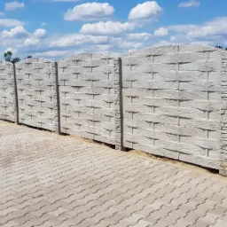 Długi, szary, betonowy płot z wzorem imitującym drewno, biegnący wzdłuż kostki brukowej pod błękitnym niebem z chmurami.