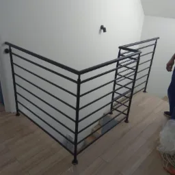 Nowoczesna, czarna poręcz schodowa z poziomymi prętami, zamontowana w jasnym wnętrzu z drewnopodobną podłogą, widok z dołu schodów, fragment sylwetki osoby po prawej stronie.