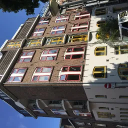 Fasada budynku z oknami o różnorodnych kolorowych ramach: czerwonych, żółtych i białych, część budynku z cegły, część biała, z zaparkowanym samochodem dostawczym w tle.