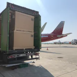 Otwarta naczepa ciężarówki z ładunkiem kartonowych pudeł na płycie lotniska, w tle samoloty z czerwonymi ogonami i oznaczeniem VQ-BSG, widoczne tablice rejestracyjne TKI 8202P