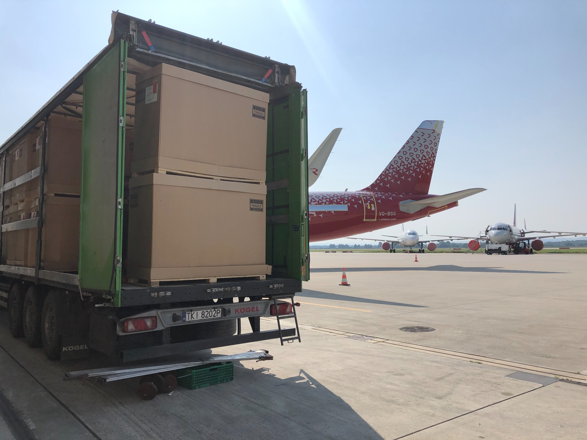 Otwarta naczepa ciężarówki z ładunkiem kartonowych pudeł na płycie lotniska, w tle samoloty z czerwonymi ogonami i oznaczeniem VQ-BSG, widoczne tablice rejestracyjne TKI 8202P