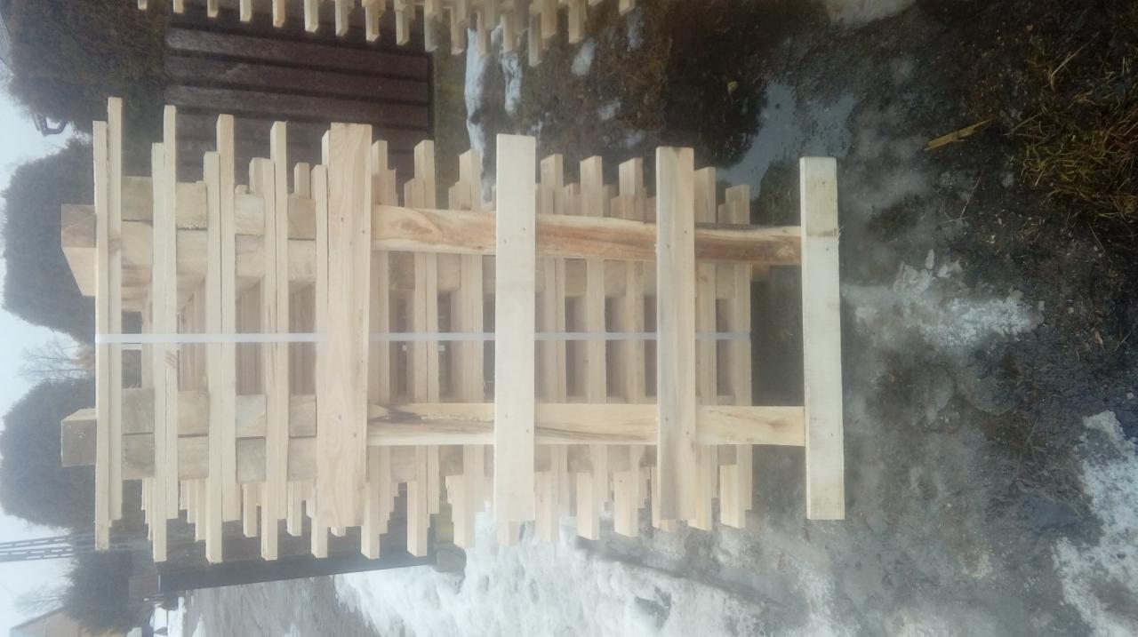 Świeżo pocięte, jasne deski ułożone w stos i zabezpieczone taśmą, widoczne na tle ziemi z resztkami śniegu.