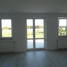 Wnętrze nowego domu z białymi ścianami, szarą podłogą imitującą panele, grzejnikami pod oknami i dużymi, przeszklonymi drzwiami balkonowymi wychodzącymi na zewnątrz.