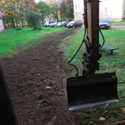 Żółty, brudny, stalowy lemiesz koparki w trakcie niwelacji terenu pokrytego trawą i świeżo przekopaną ziemią, w tle budynki mieszkalne i zaparkowane samochody.