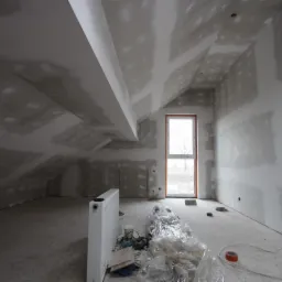 Poddasze w trakcie wykańczania płytami kartonowo-gipsowymi, widoczne szpachlowane łączenia, okno z widokiem na drzewo, grzejnik i narzędzia budowlane na podłodze.