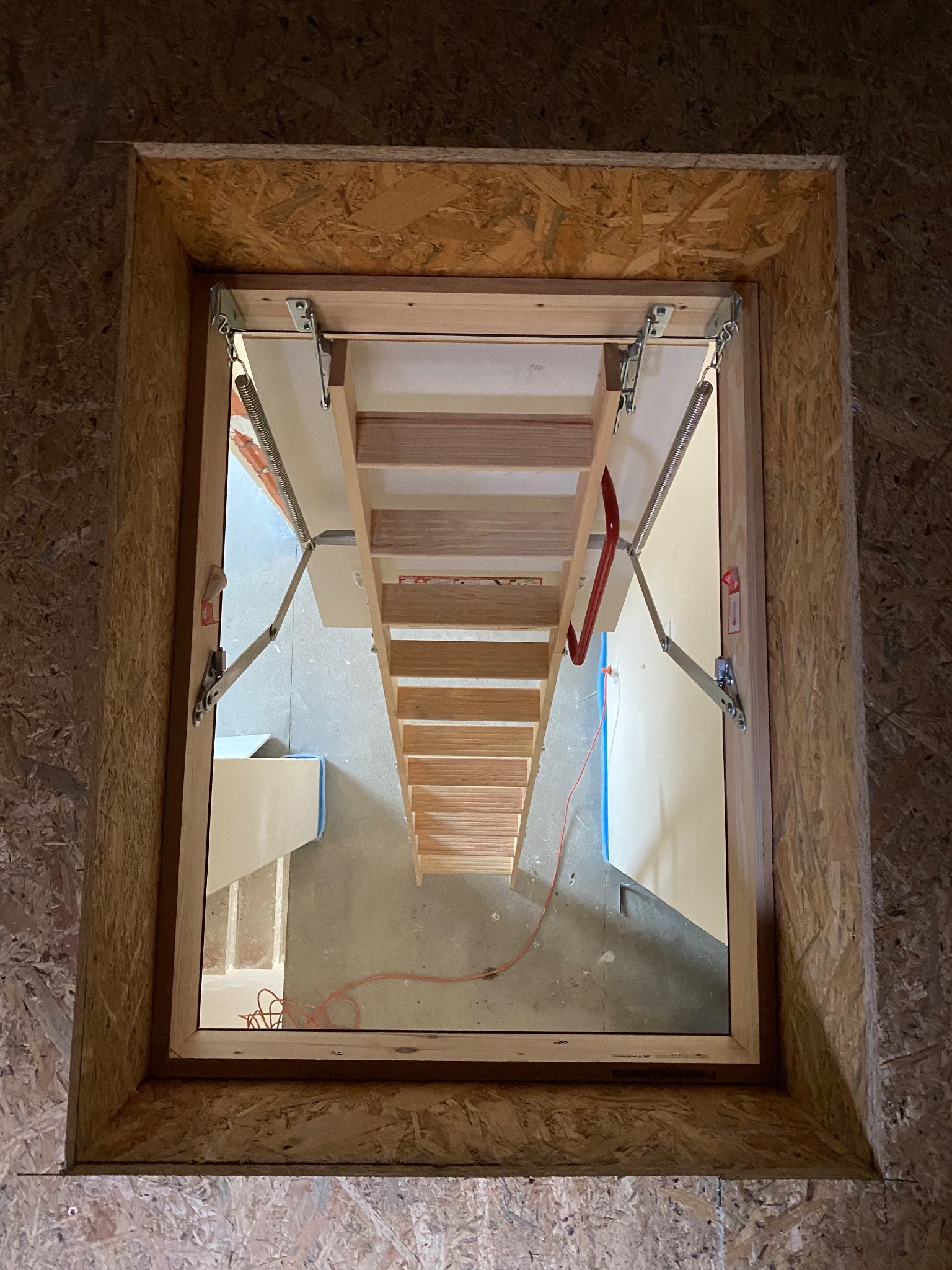 Zamontowane drewniane schody strychowe, widok z góry na otwarty właz w poddaszu wykończonym płytą OSB. Widoczne elementy mechanizmu składania schodów.