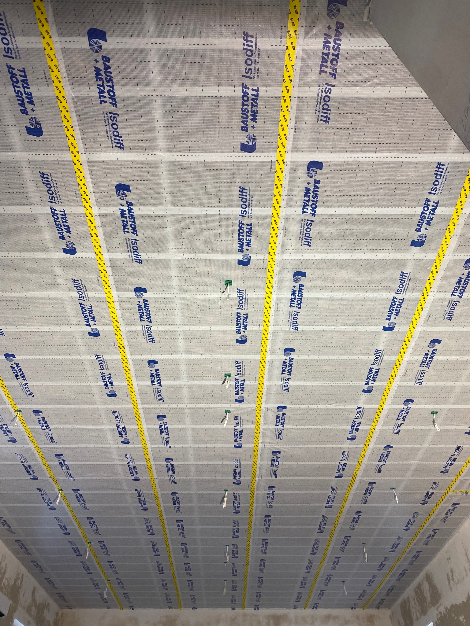 Montaż folii paroizolacyjnej Baustoff Isodiff z metalem, zabezpieczonej żółtą taśmą, na suficie. Widoczne zszywki mocujące i fragment ściany w tle.