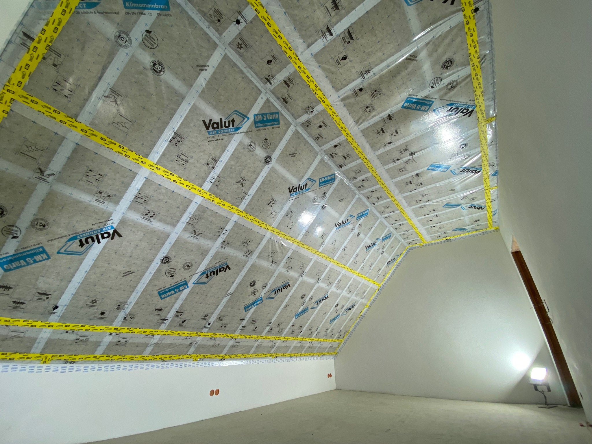 Poddasze z folią paroizolacyjną Valut KM-5 Vario, zabezpieczoną żółtą taśmą. Widoczne detale montażu i struktura membrany. Oświetlenie robocze w rogu pomieszczenia.