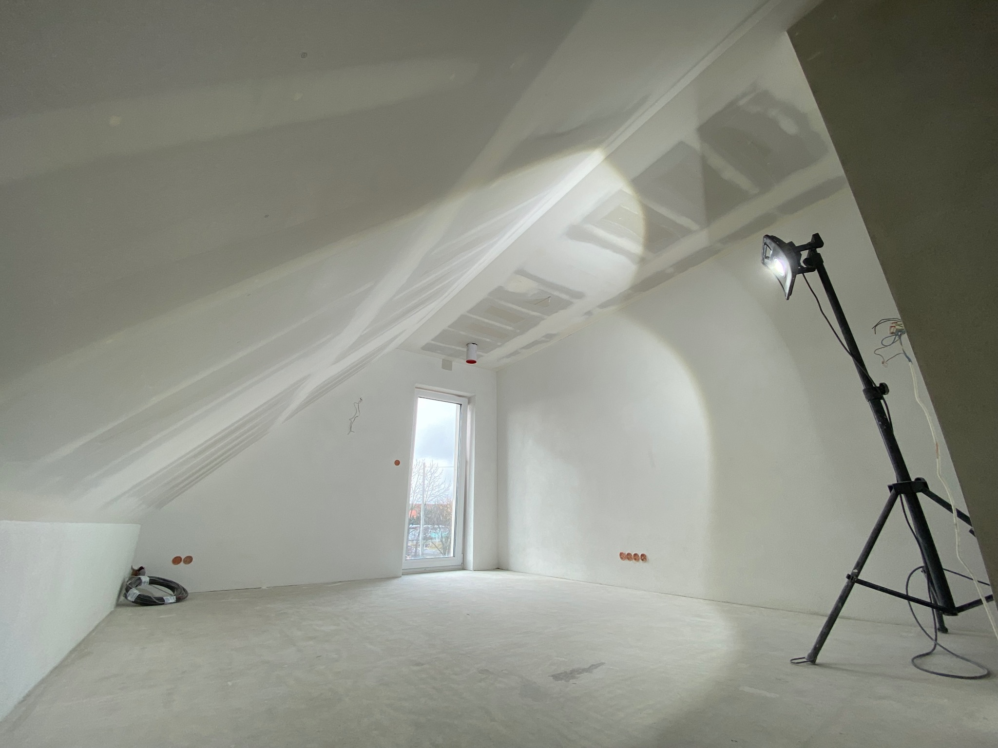 Poddasze po montażu płyt gipsowo-kartonowych, widoczne szpachlowane łączenia, oświetlenie budowlane i okno. Przygotowanie do malowania.