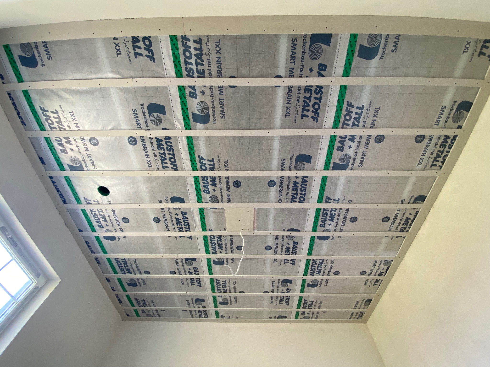 Montaż izolacji poddasza z folią paroizolacyjną z nadrukiem, widoczne drewniane belki i fragment okna dachowego. Prace wykończeniowe w trakcie realizacji.