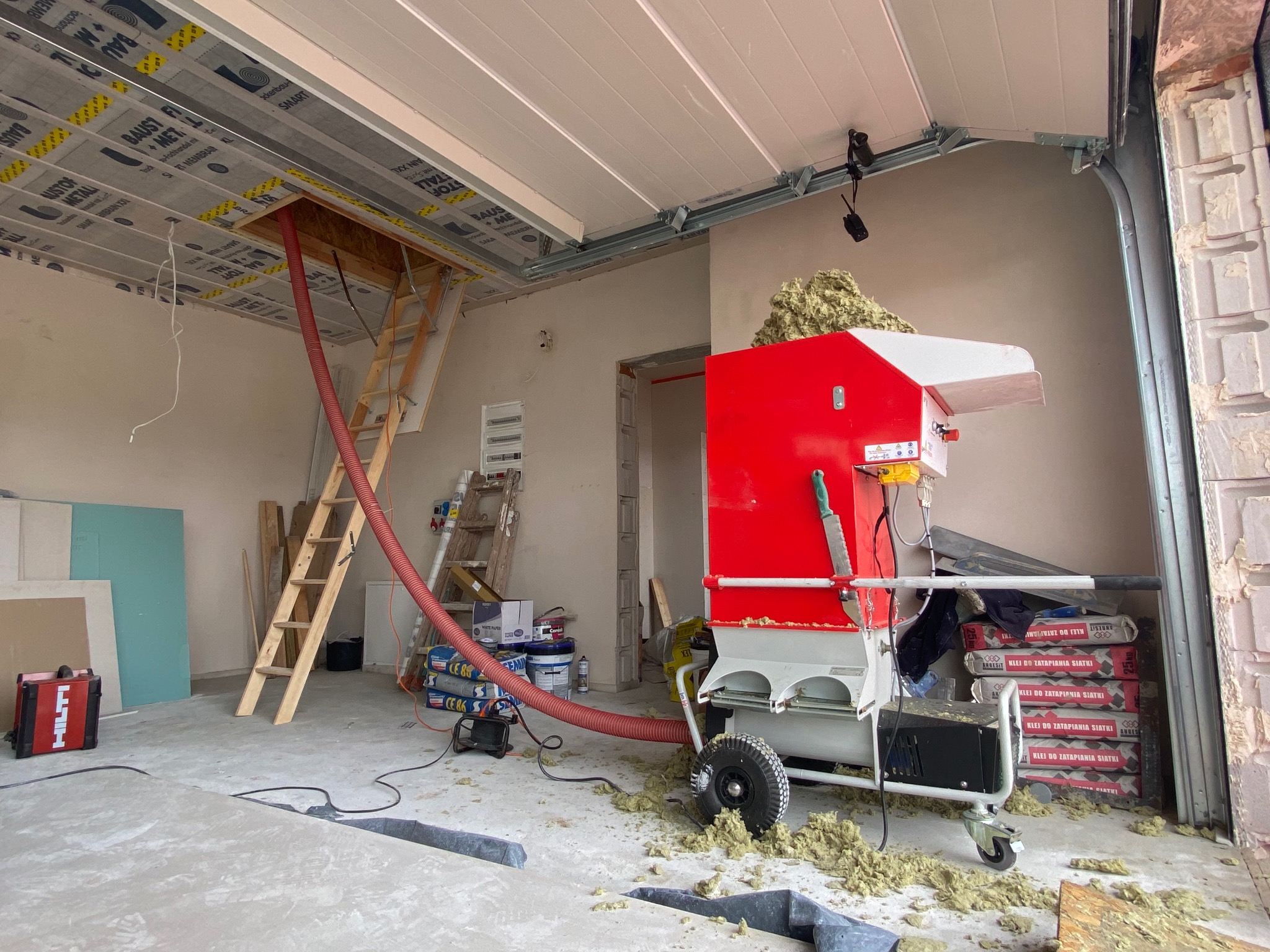 Maszyna do wdmuchiwania izolacji w trakcie pracy na poddaszu. Widoczny wąż doprowadzający materiał izolacyjny i rozłożone materiały budowlane.