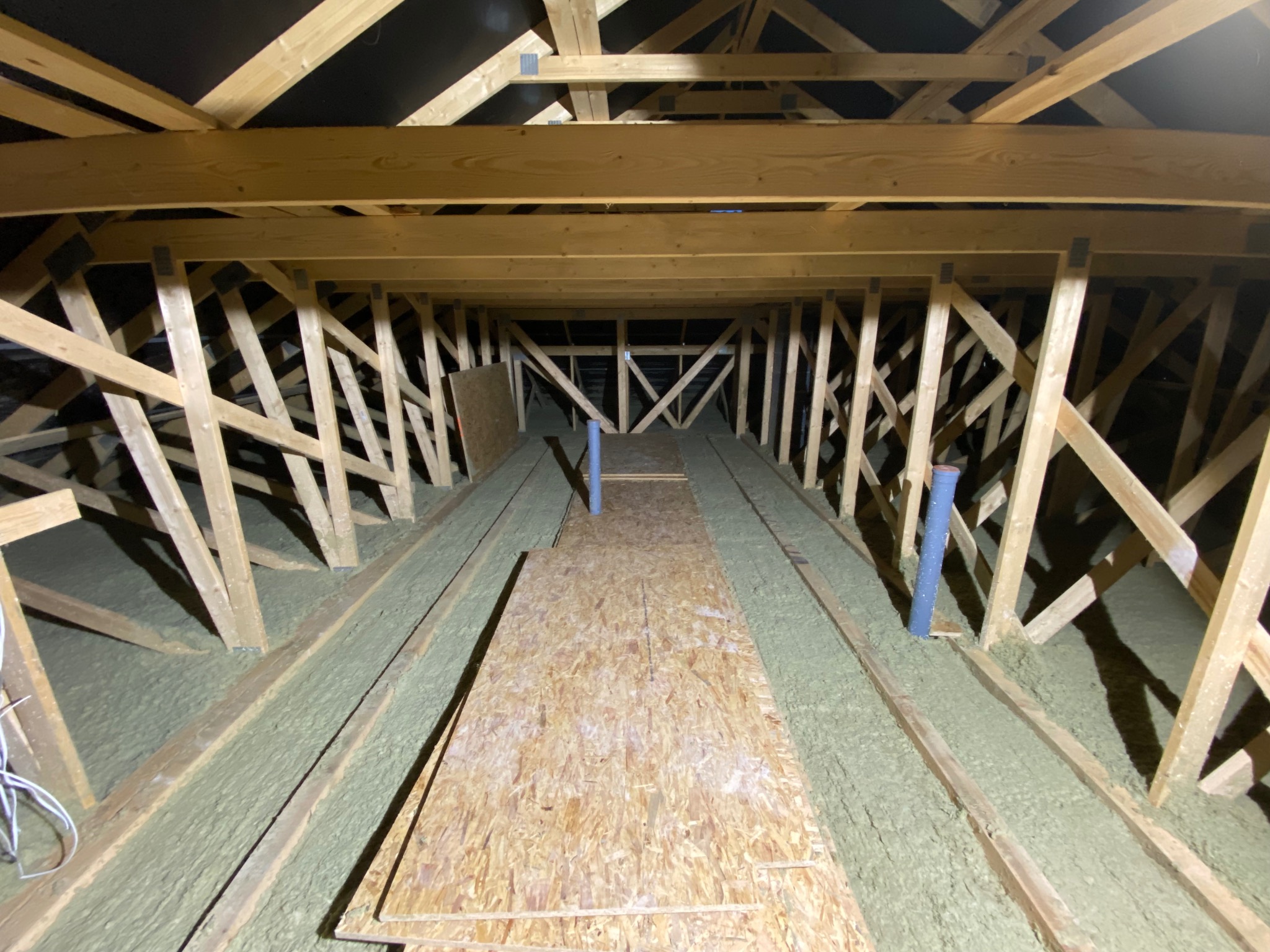 Poddasze z drewnianą konstrukcją, ocieplone wełną mineralną, z fragmentami płyt OSB. Widoczne rury wentylacyjne. Prace izolacyjne w trakcie realizacji.