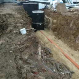 Wykop pod instalację przydomowej oczyszczalni ścieków z widocznymi betonowymi kręgami i rurą kanalizacyjną w ziemi, narzędzia na ziemi, budynek w tle.