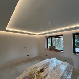 Firma DAR-GOŚ Dariusz Nowak - Pokój w trakcie wykończenia: surowa podłoga, ściany w kolorze złamanej bieli, podwieszany sufit z oświetleniem LED po obwodzie, okna z widokiem na zieleń oraz materiały budowlane zabezpieczone...