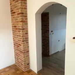 Wnętrze z dekoracyjnym narożnikiem z cegły klinkierowej, widoczne parkiet drewniany z ciemnymi akcentami i łukowate przejście do kolejnego pomieszczenia z drewnianą podłogą i surowymi ścianami.