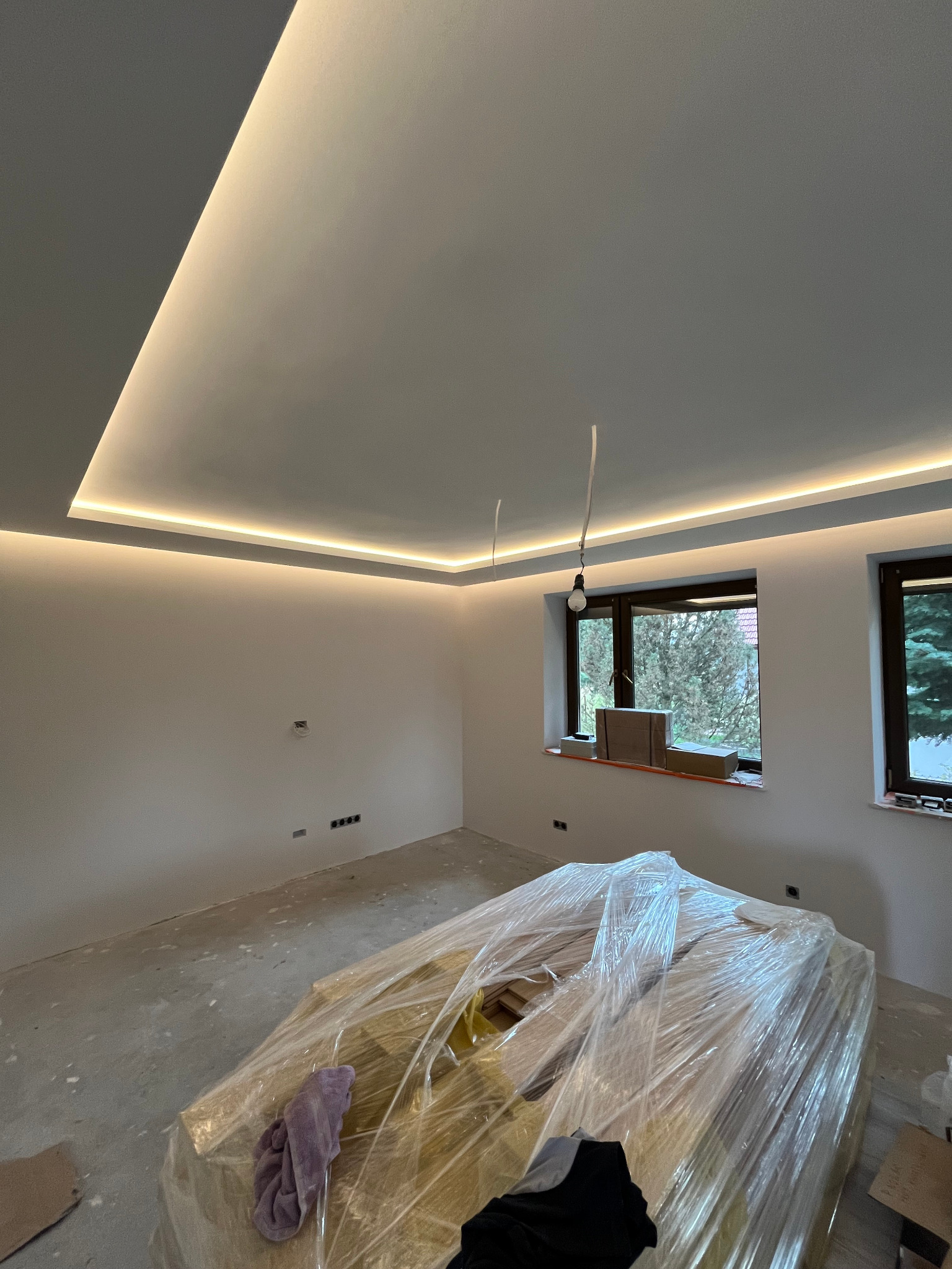 Pokój w trakcie wykończenia: surowa podłoga, ściany w kolorze złamanej bieli, podwieszany sufit z oświetleniem LED po obwodzie, okna z widokiem na zieleń oraz materiały budowlane zabezpieczone...