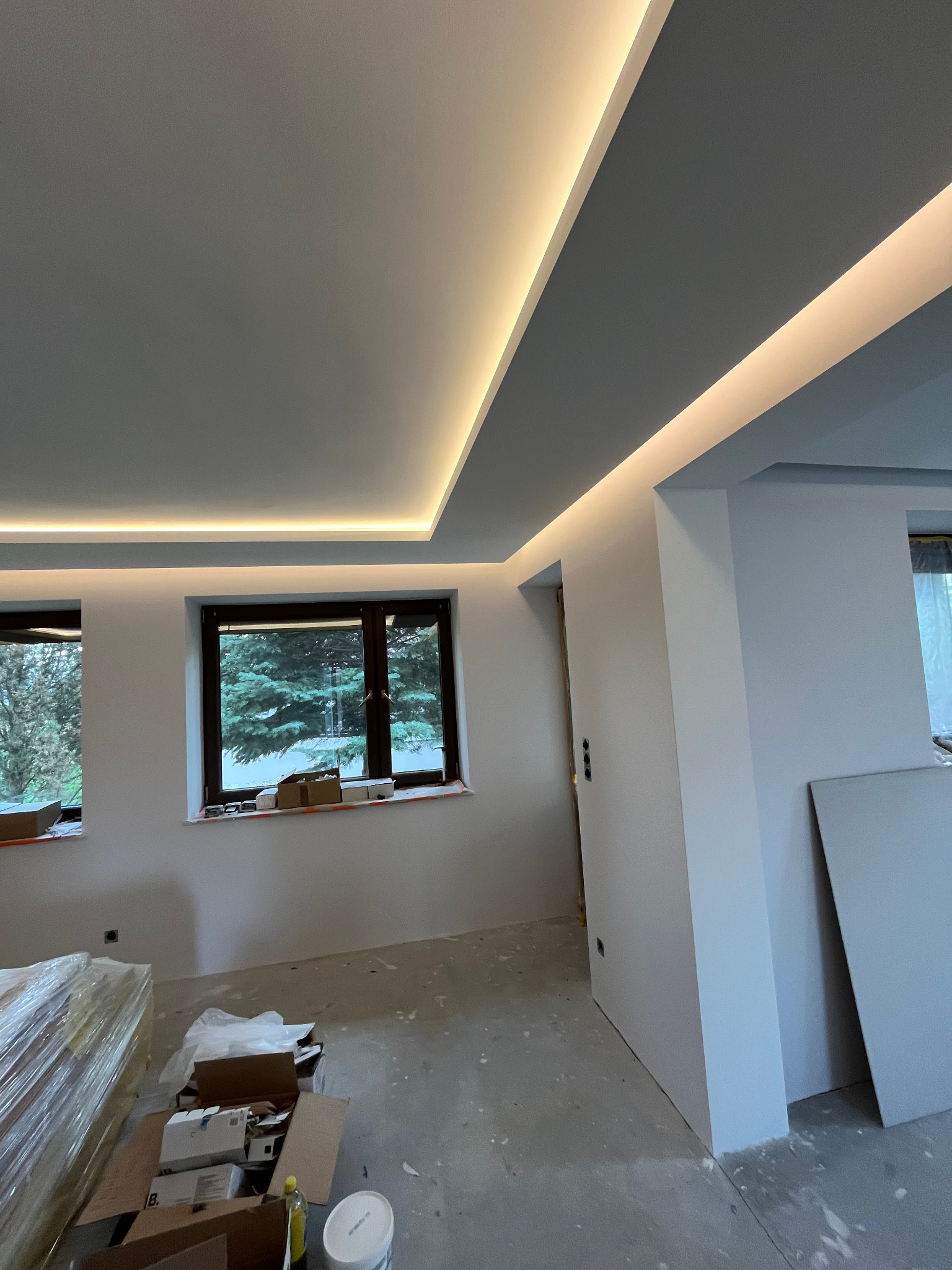 Wnętrze w trakcie prac wykończeniowych: sufit podwieszany z oświetleniem LED, dwa okna z widokiem na zewnątrz, na podłodze kartony i materiały budowlane.