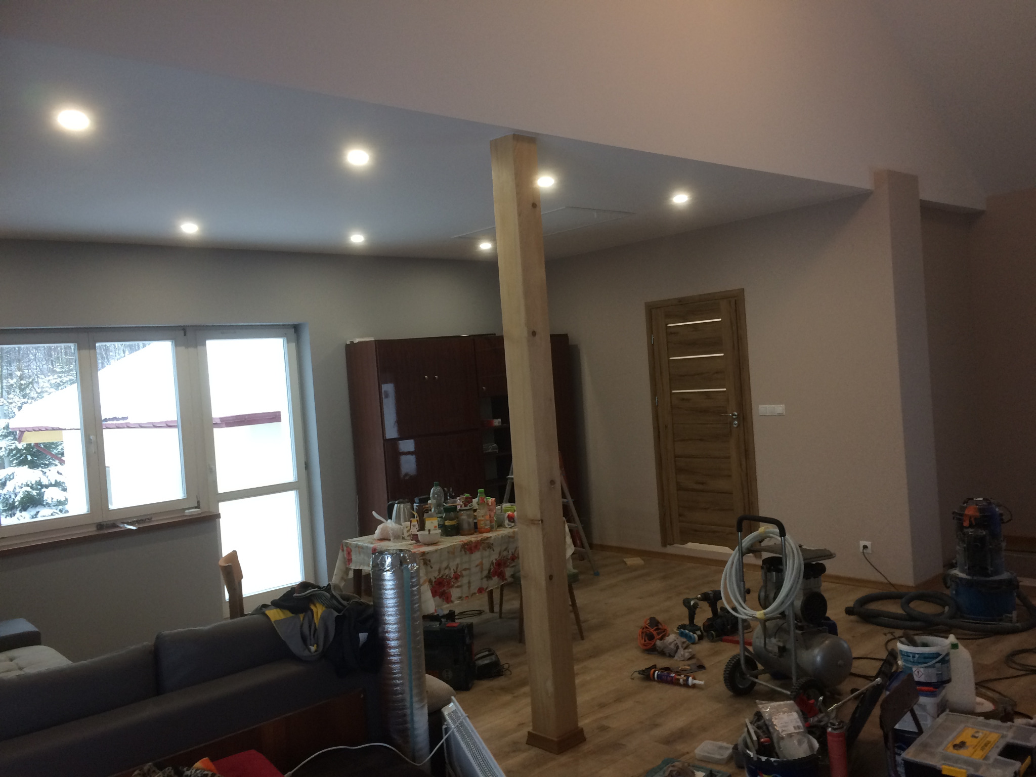 Wnętrze w trakcie prac wykończeniowych: widoczny słup konstrukcyjny, narzędzia, kompresor, stół z artykułami spożywczymi, okno z widokiem na zaśnieżony ogród.