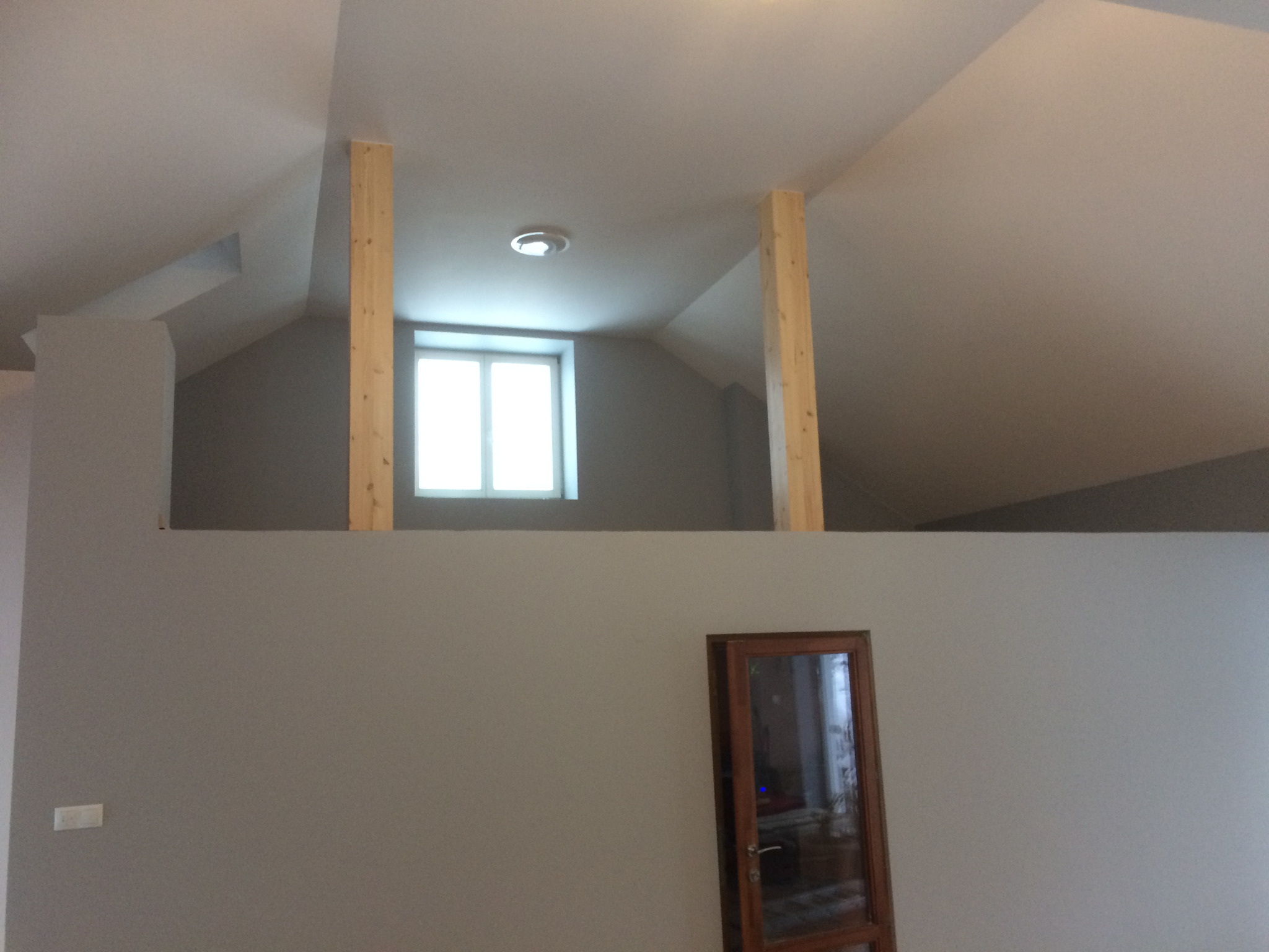 Wnętrze poddasza z drewnianymi słupami podtrzymującymi strop, widoczne okno na górze i przeszklone drzwi na dole, ściany pomalowane na szaro.
