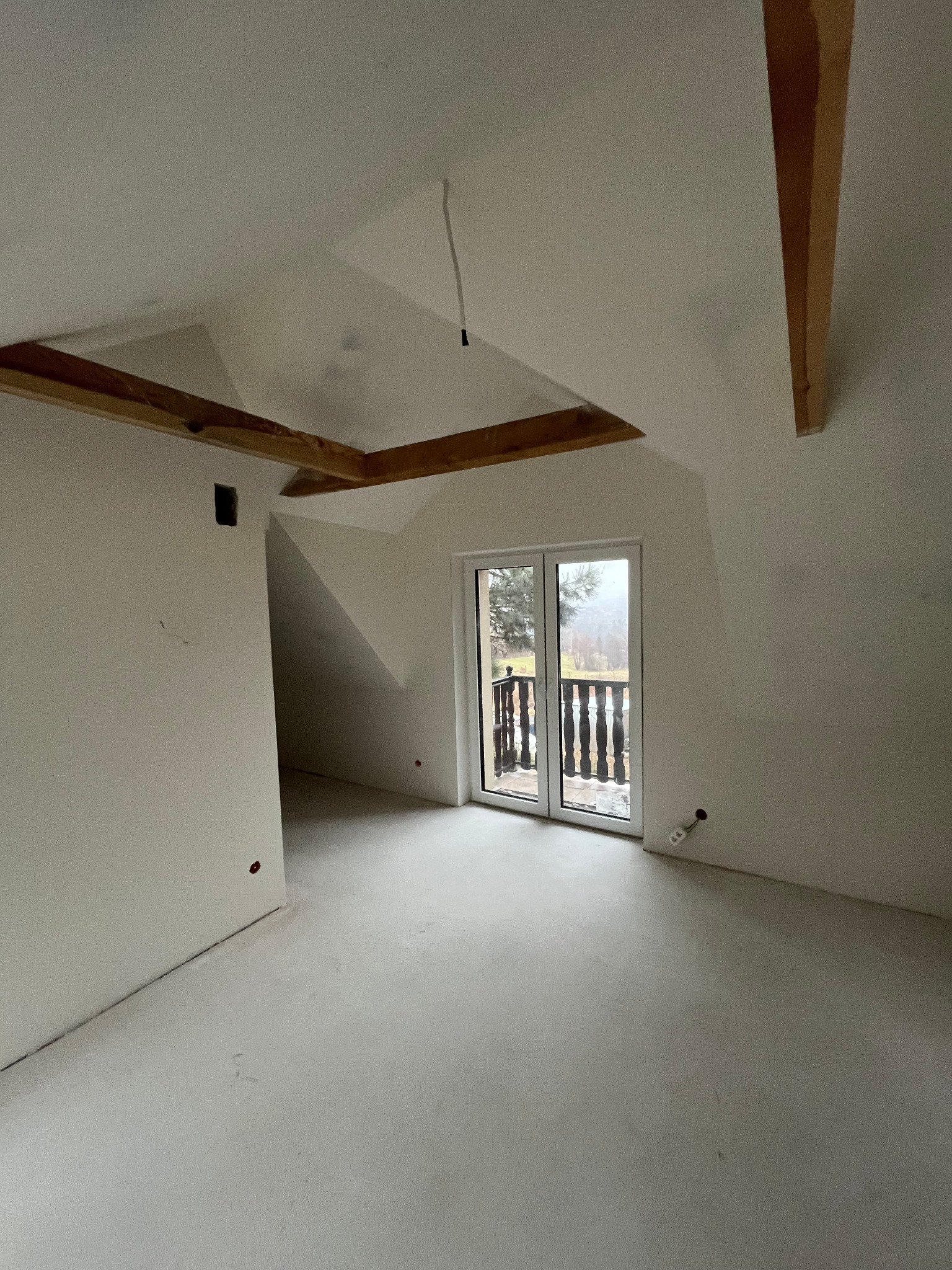 Poddasze w stanie deweloperskim z surowymi belkami stropowymi, białymi ścianami i wylewką, okno balkonowe z widokiem na zewnątrz.