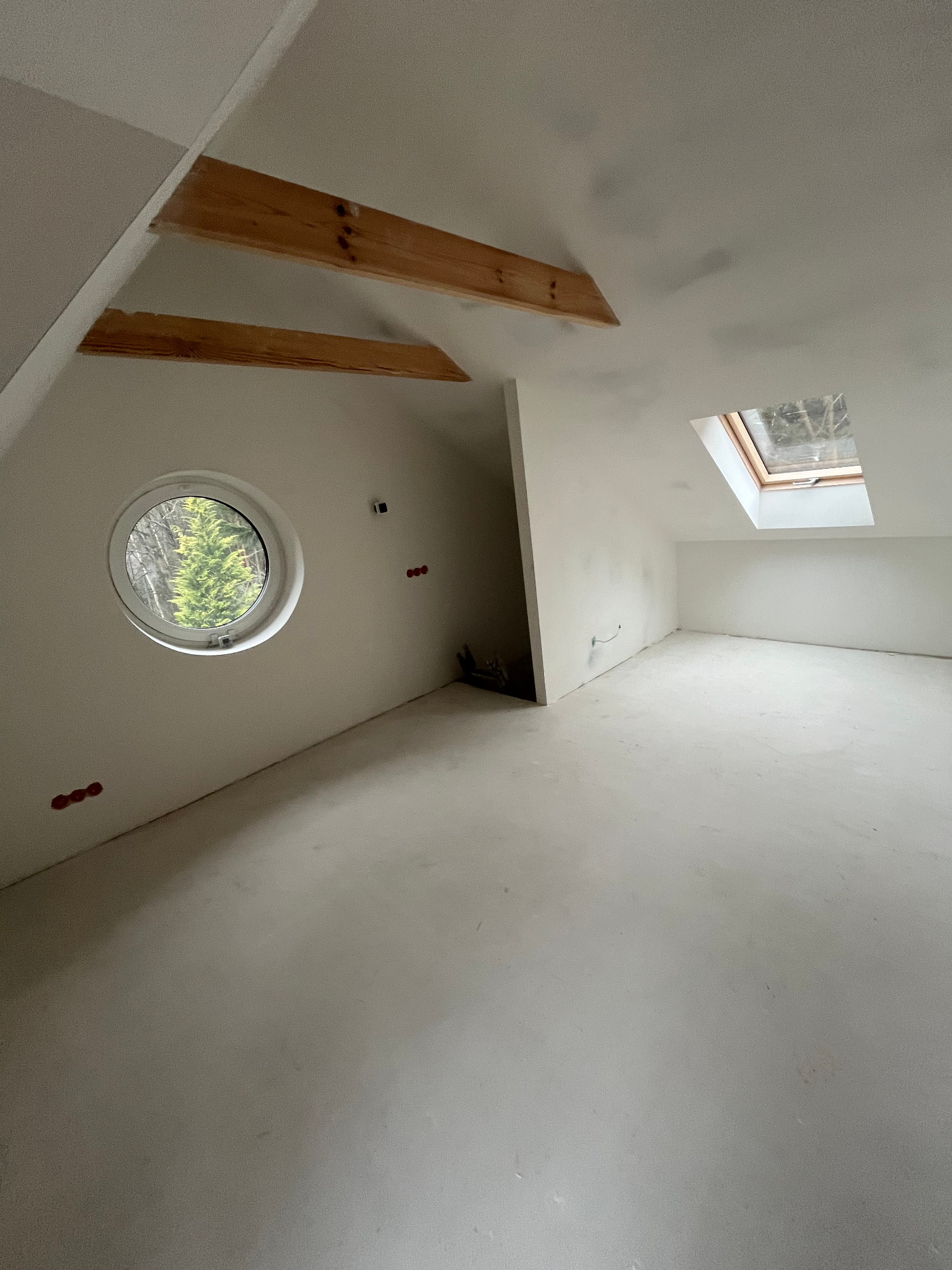 Poddasze w stanie deweloperskim z oknem dachowym i okrągłym oknem z widokiem na drzewa, widoczne drewniane elementy konstrukcyjne poddasza, ściany przygotowane do malowania z wystającymi przewodami...