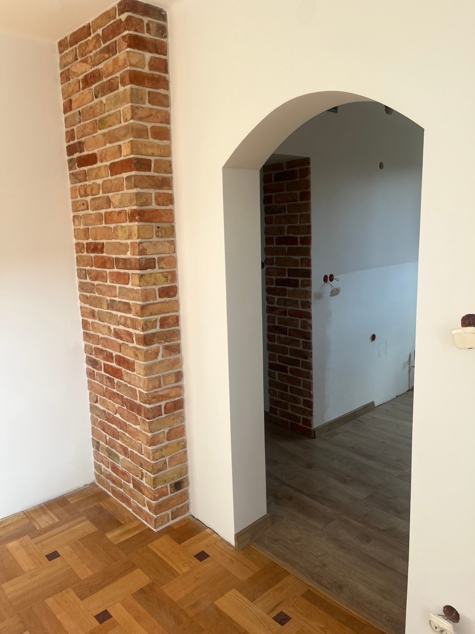 Wnętrze z dekoracyjnym narożnikiem z cegły klinkierowej, widoczne parkiet drewniany z ciemnymi akcentami i łukowate przejście do kolejnego pomieszczenia z drewnianą podłogą i surowymi ścianami.