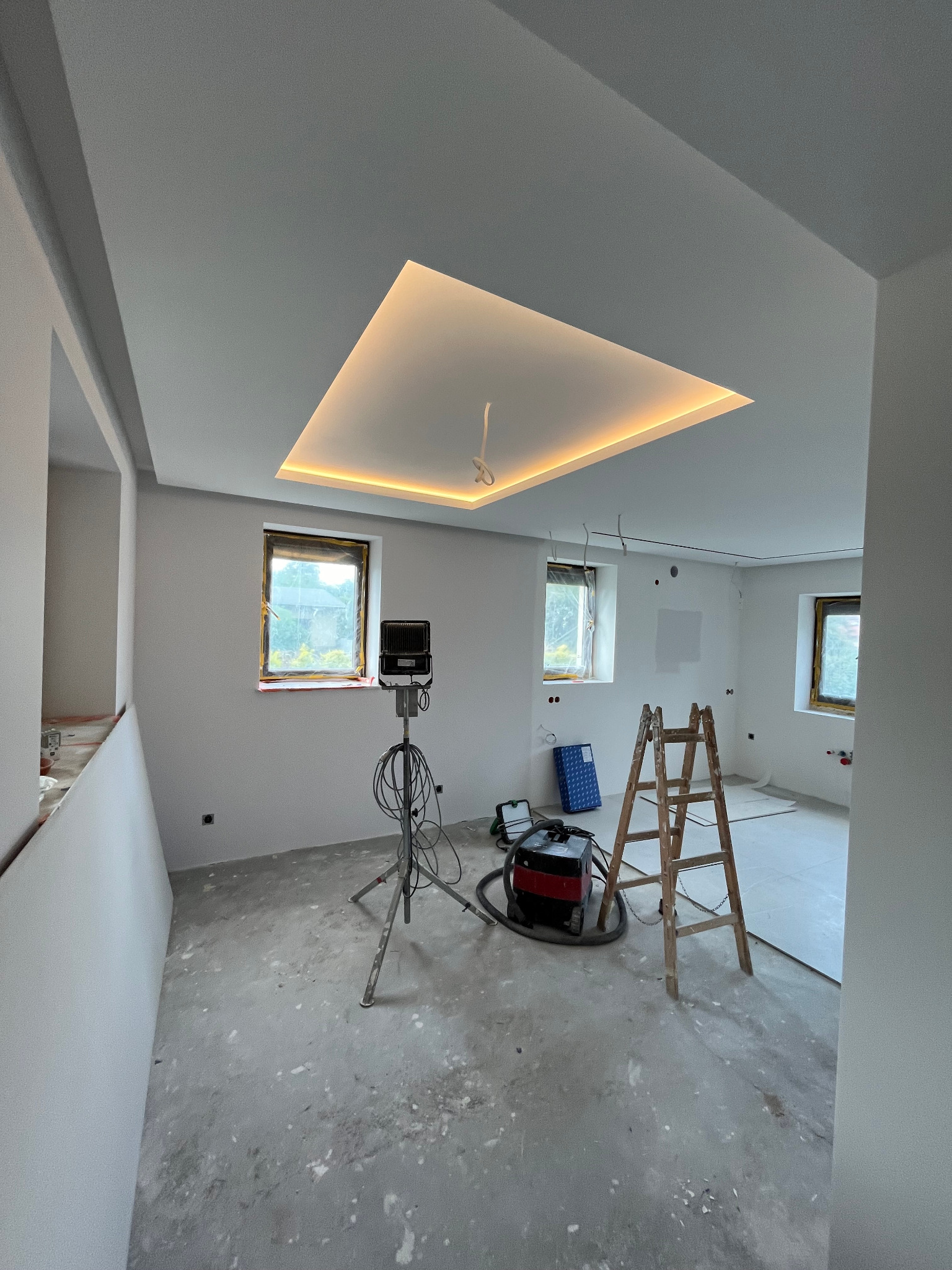 Wnętrze w trakcie wykańczania: widoczny sufit z podświetleniem LED w kształcie prostokąta, drabina, lampa budowlana na statywie oraz odkurzacz przemysłowy, okna zabezpieczone folią.
