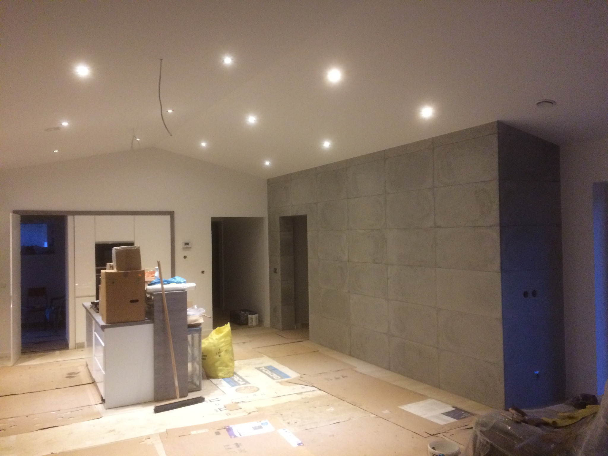 Wnętrze w trakcie prac wykończeniowych: ściana wyłożona płytami imitującymi beton, kartony i materiały budowlane na podłodze, widoczna instalacja elektryczna pod sufitem.