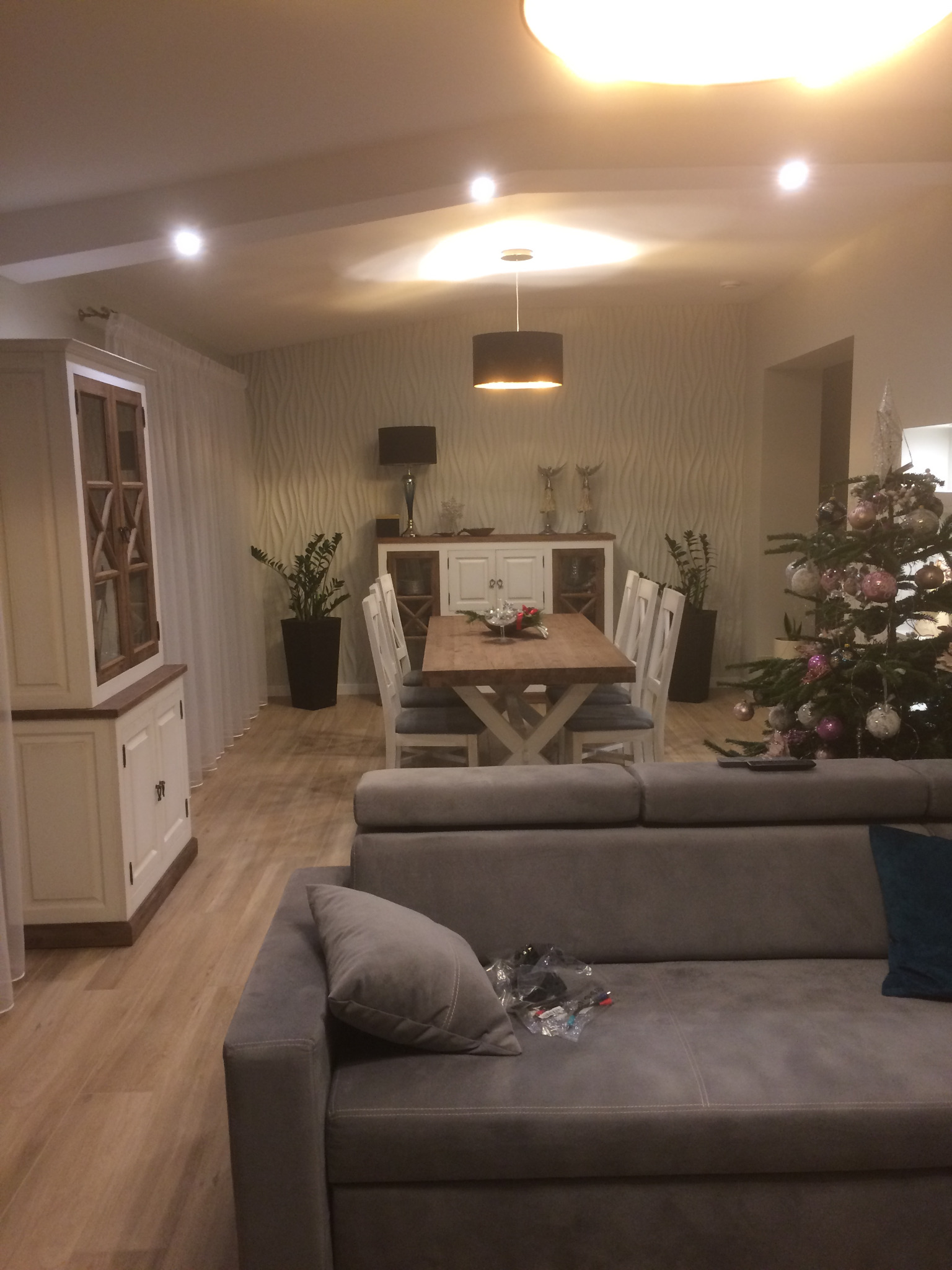 Jasne wnętrze salonu z jadalnią, drewniany stół z białymi krzesłami, szara sofa z poduszkami, biała komoda i kredens, choinka z ozdobami oraz rośliny doniczkowe.