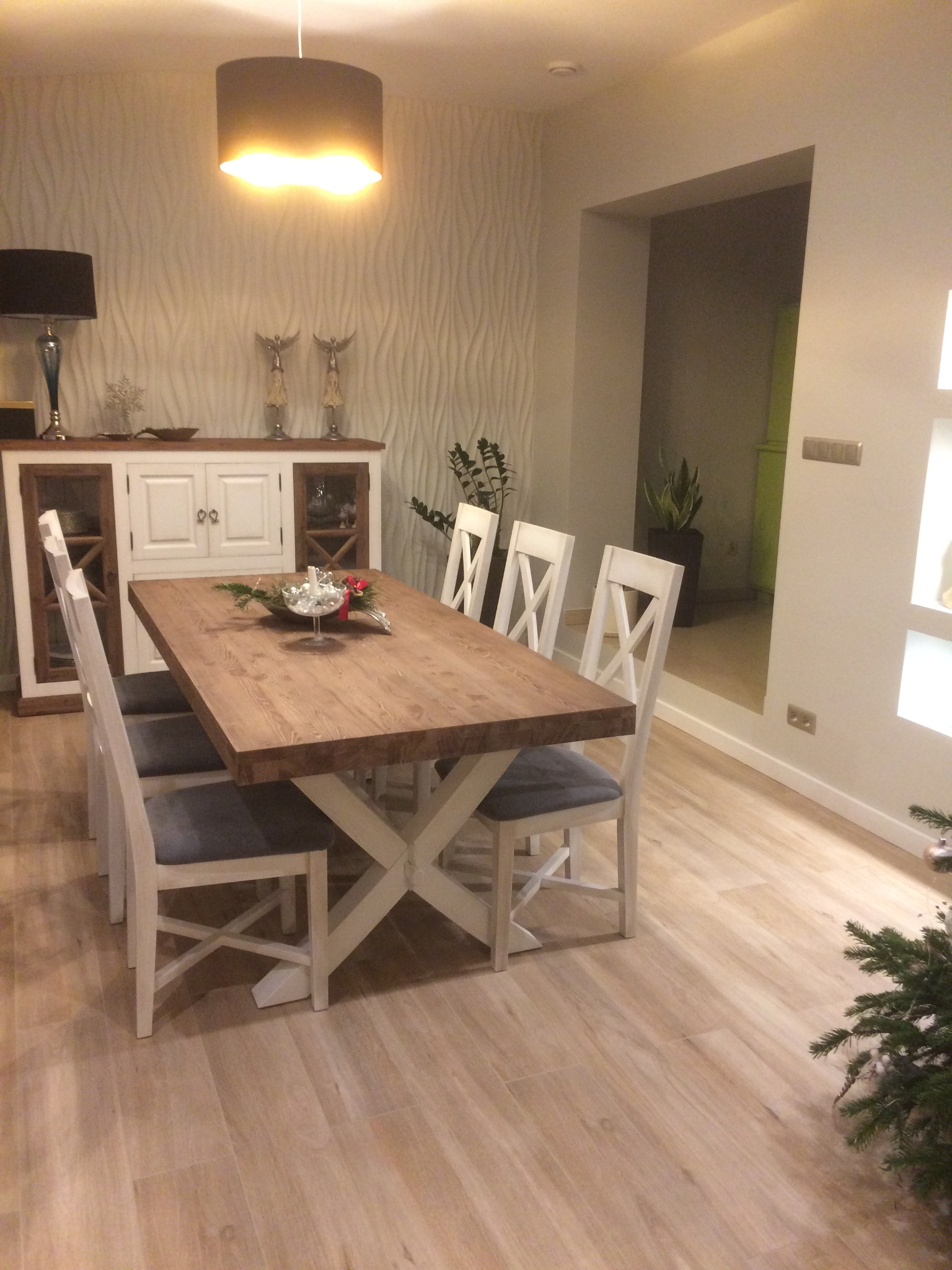 Jasne wnętrze jadalni z drewnianym stołem na białej nodze w kształcie litery X, białymi krzesłami z szarymi poduszkami i komodą w tle na tle ściany z dekoracyjną fakturą.