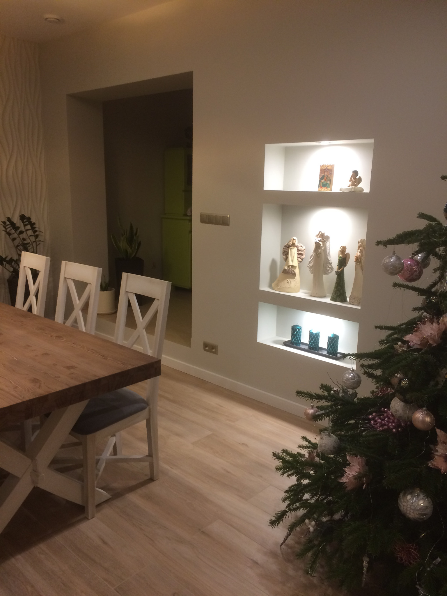 Jadalnia z drewnianym stołem i białymi krzesłami, podświetlane nisze w ścianie z dekoracjami świątecznymi, w tle widoczna zielona lodówka.