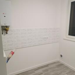 Usługi Ogólno Budowlane - Wnętrze pomieszczenia po remoncie: szare płytki imitujące drewno na podłodze, białe ściany z dekoracyjnymi płytkami nad gniazdkami elektrycznymi, piec gazowy na ścianie, białe okno.