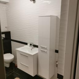 Usługi Ogólno Budowlane - Nowoczesna łazienka z białymi, falistymi płytkami na ścianach, czarną listwą dekoracyjną, czarną podłogą z białymi żyłami, białą szafką pod umywalką i wysoką białą szafką łazienkową