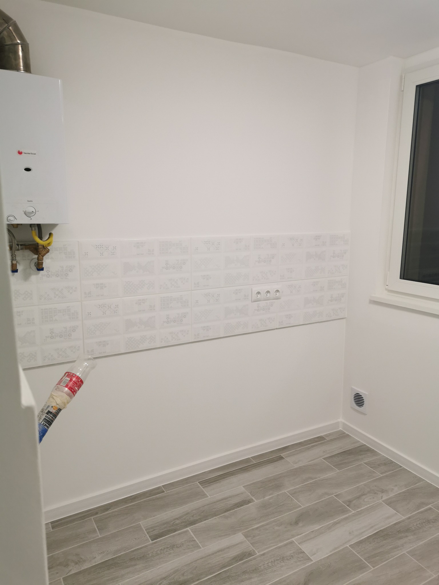 Wnętrze pomieszczenia po remoncie: szare płytki imitujące drewno na podłodze, białe ściany z dekoracyjnymi płytkami nad gniazdkami elektrycznymi, piec gazowy na ścianie, białe okno.